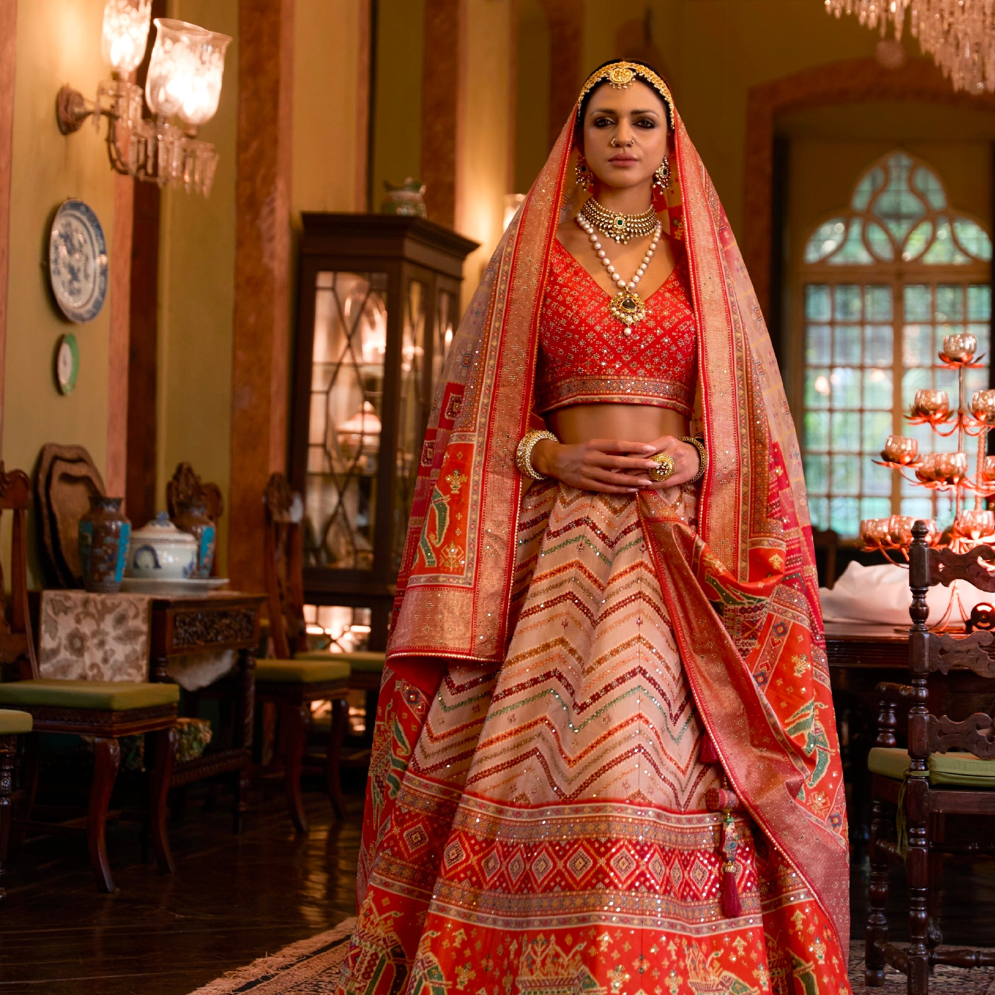 A woman wearing a traditional Indian wedding lehenga choli with mirror work, in a richly decorated room with elegant furnishings.