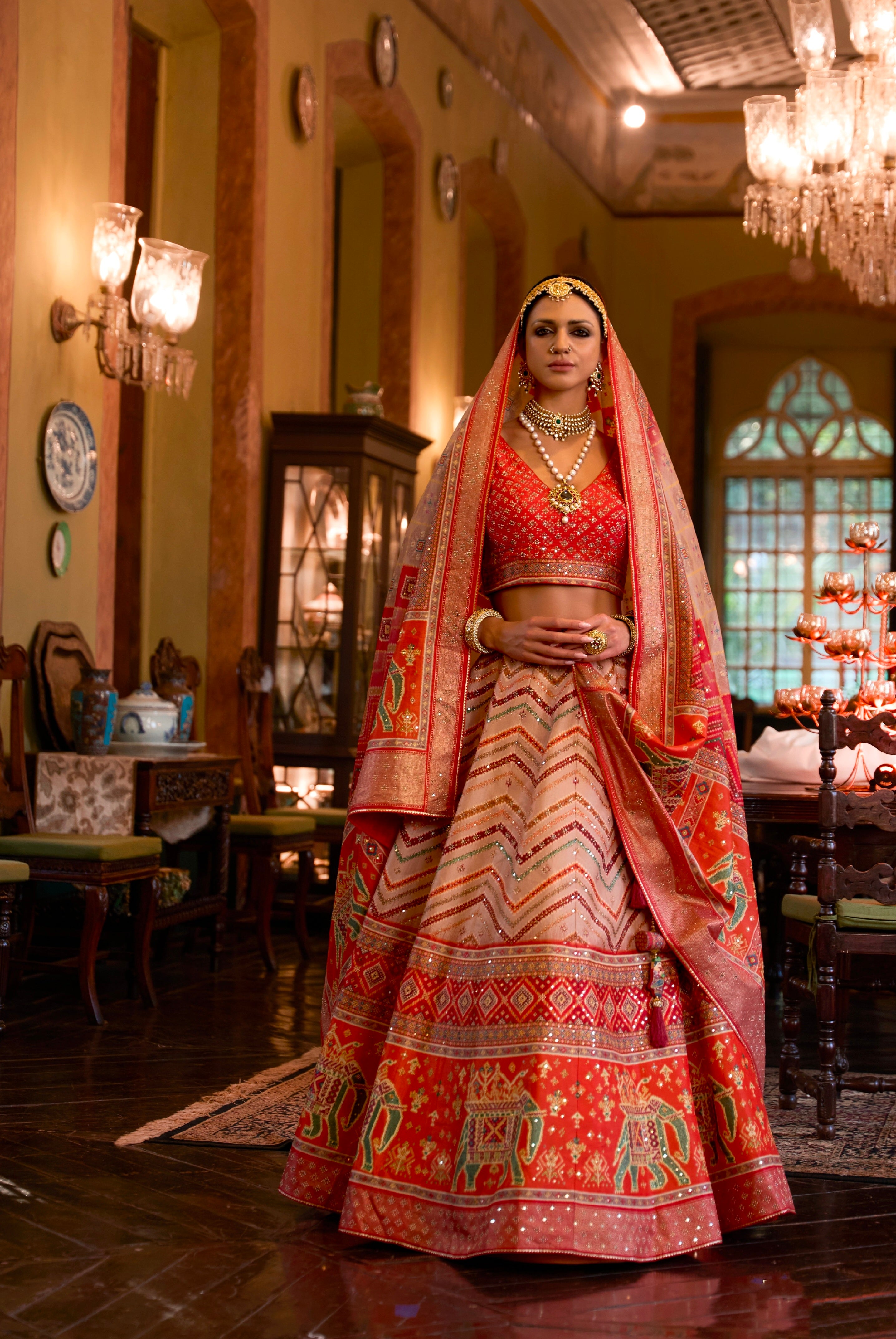 A woman wearing a traditional Indian wedding lehenga choli with mirror work, in a richly decorated room with elegant furnishings.